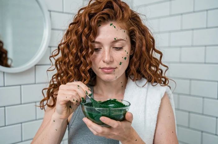 Femme aux cheveux bouclés appliquant un masque à la spiruline verte directement sur sa chevelure dans une salle de bain épurée